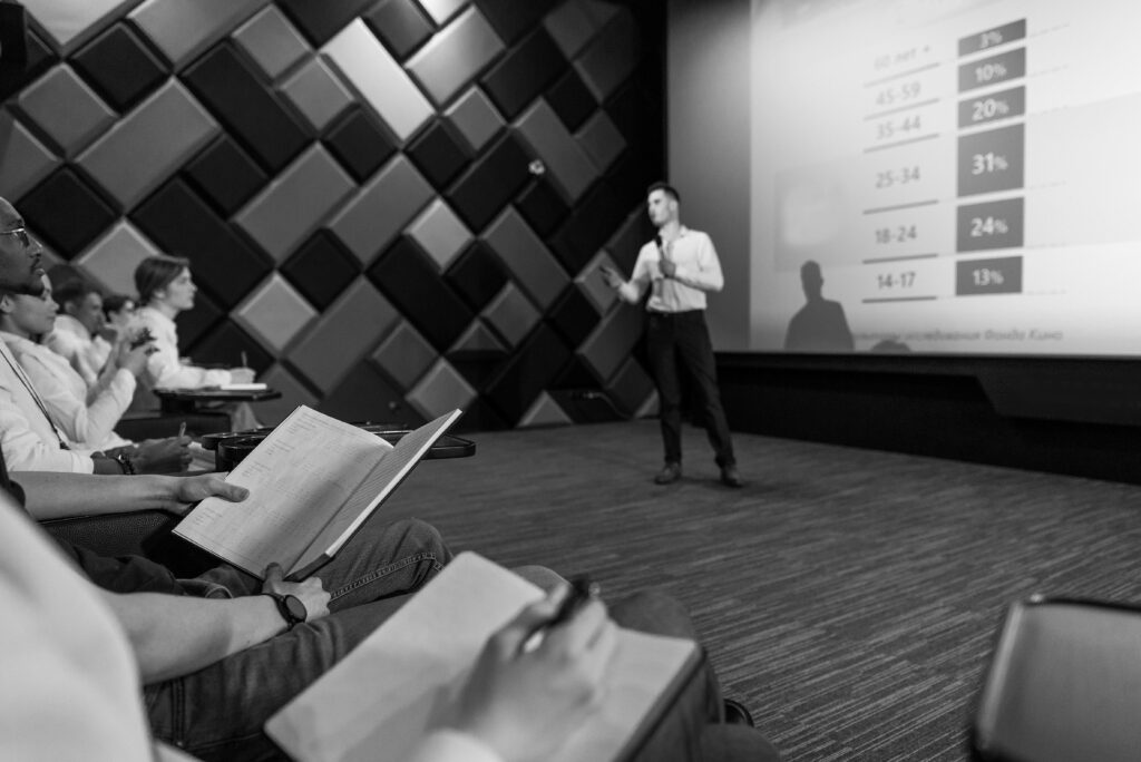 Monochrome image of a business presentation with an audience taking notes.